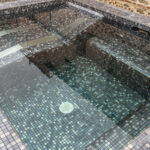 Een warm buitenzwembad met glasmozaïek in een mix van lichtgrijs en donkergrijs matjes